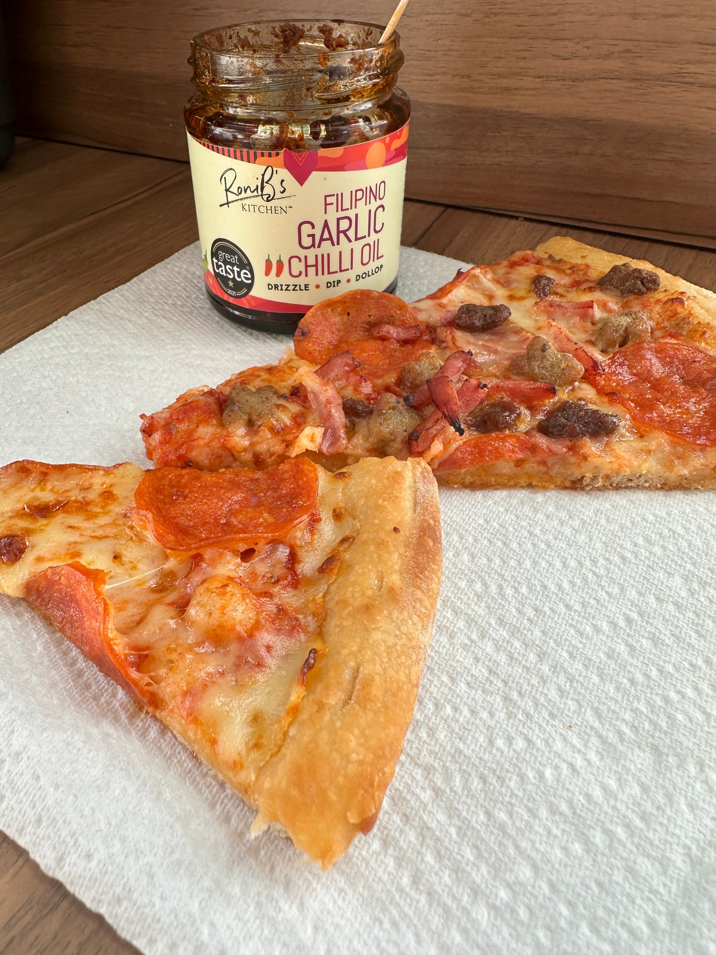 Two slices of pizza on a white napkin with an opened jar of RoniB's Kitchen Garlic Chilli Oil with a teaspoon inside the jar.