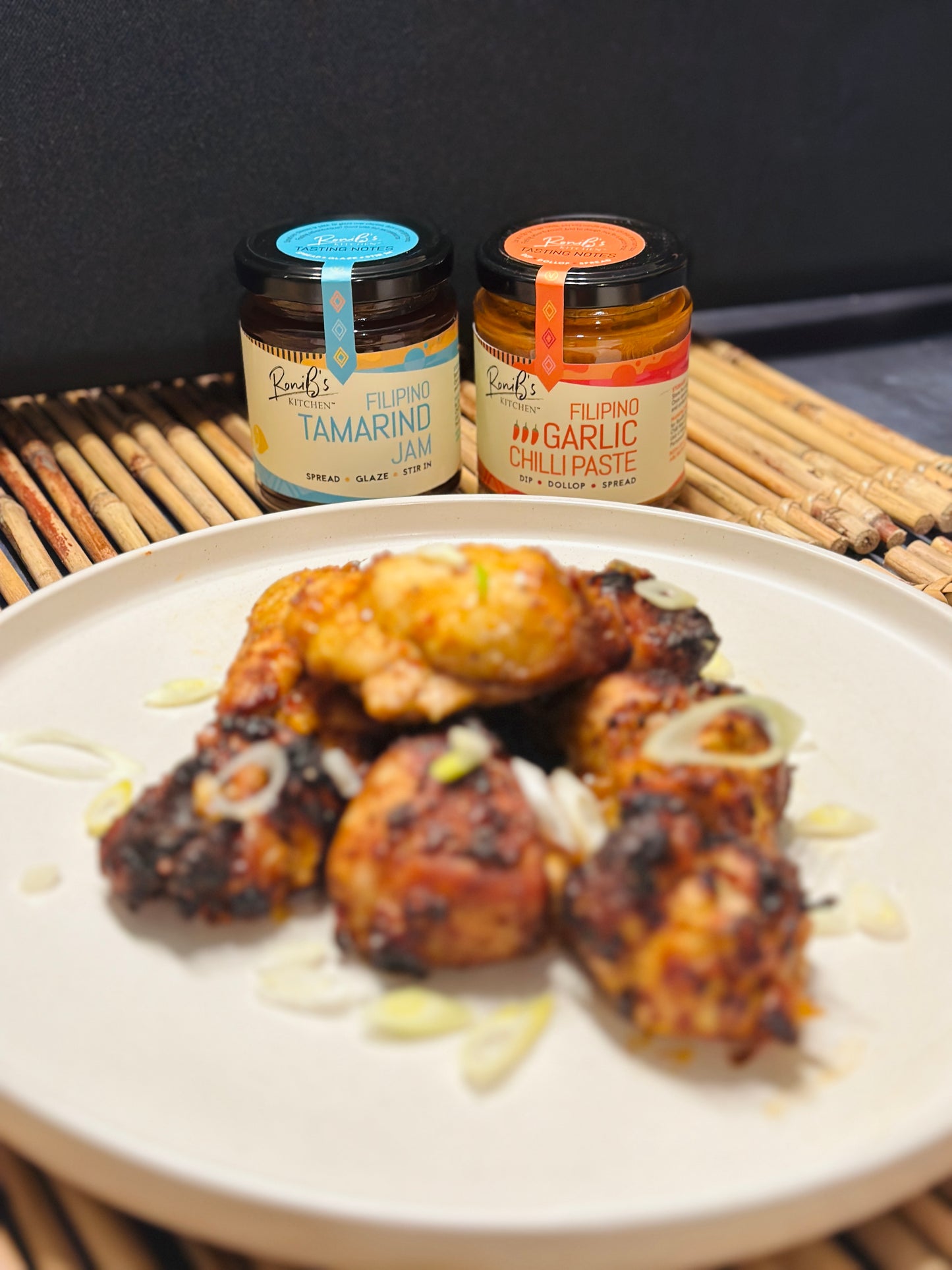 Chunks of chicken topped with thinly sliced spring onions served on a white round plate with RoniB's Kitchen Garlic Chilli Paste and Tamarind Jam in the background.