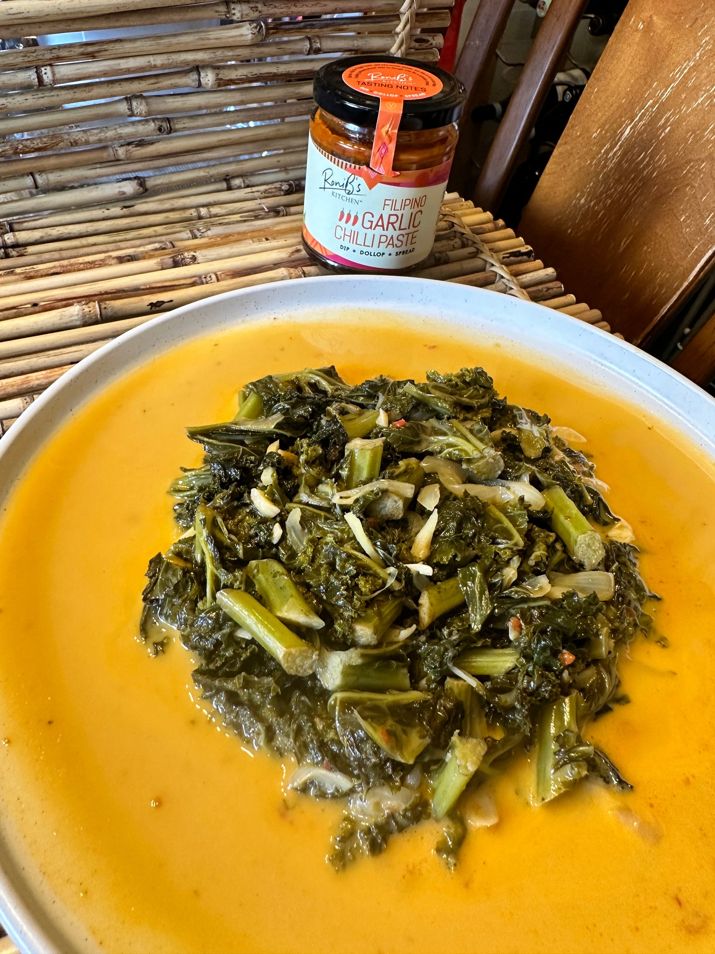 A plate of kale laing dish served in a white round plate on a bamboo with placemat with RoniB's Kitchen Garlic Chilli Paste in the background.