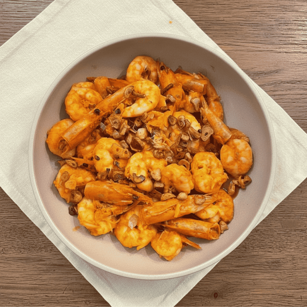 Close-up of Filipino-style gambas featuring orange prawns in a glossy garlic chilli sauce, topped with crispy toasted garlic and prawn heads, served in a neutral bowl on a wooden table.