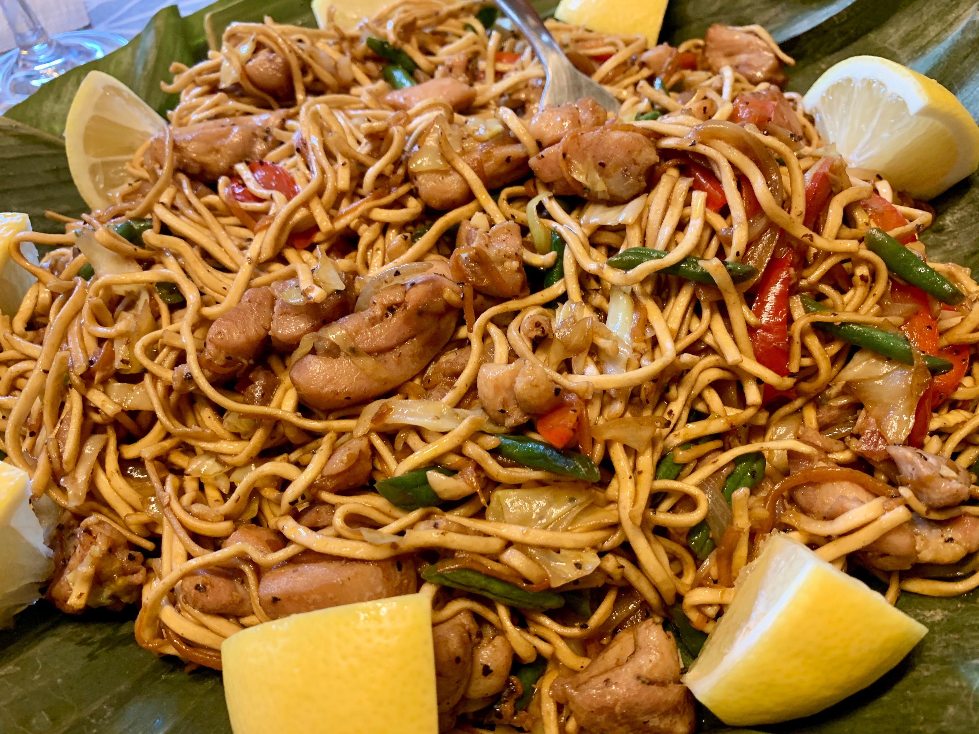 close up shot of pancit canton served in a bilao lined with banana leaves. slices of lemon on the side of the noodles.
