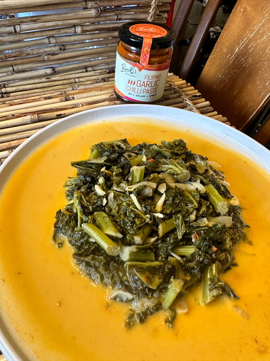 A plate of kale laing dish served in a white round plate on a bamboo with placemat with RoniB's Kitchen Garlic Chilli Paste in the background.