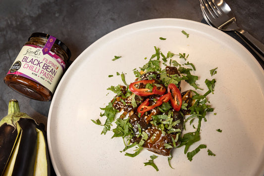 A top view of aubergine dish cooked with RoniB's Kitchen Black bean chilli paste served on a white round plate, spoon and fork on the top right hand side, a jar of RoniB's Kitchen black bean chilli paste on the left side of the plate and a sliced fresh aubergine in the lower left hand side of the plate.