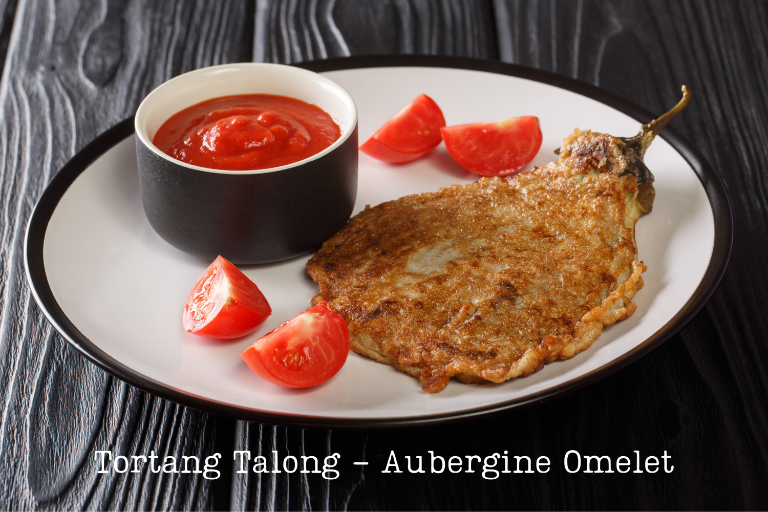 aubergine omelette on a white place with banana ketchup and sliced tomatoes on a black table top