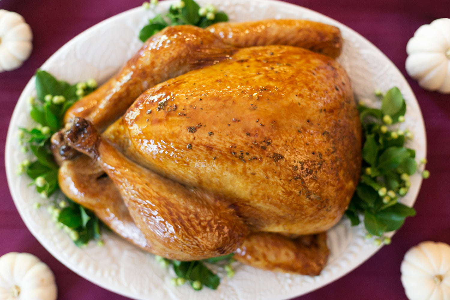 A white plate with a whole roasted turkey with leaves and flowers as dish decorations on a table with red table cloth and garlic bulbs on all four sides